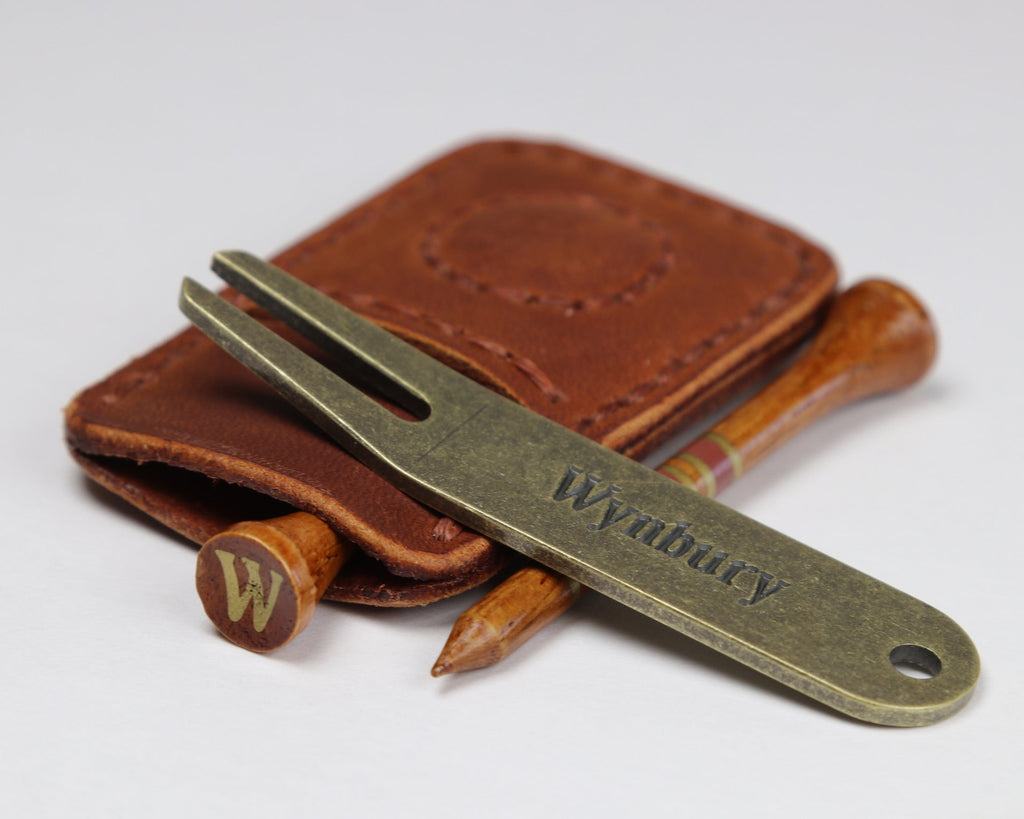 A metal golf divot tool with the engraved name 'Wynbury' resting on a chestnut brown full grain leather pouch, with two wooden golf tees visible behind it on a light gray background.